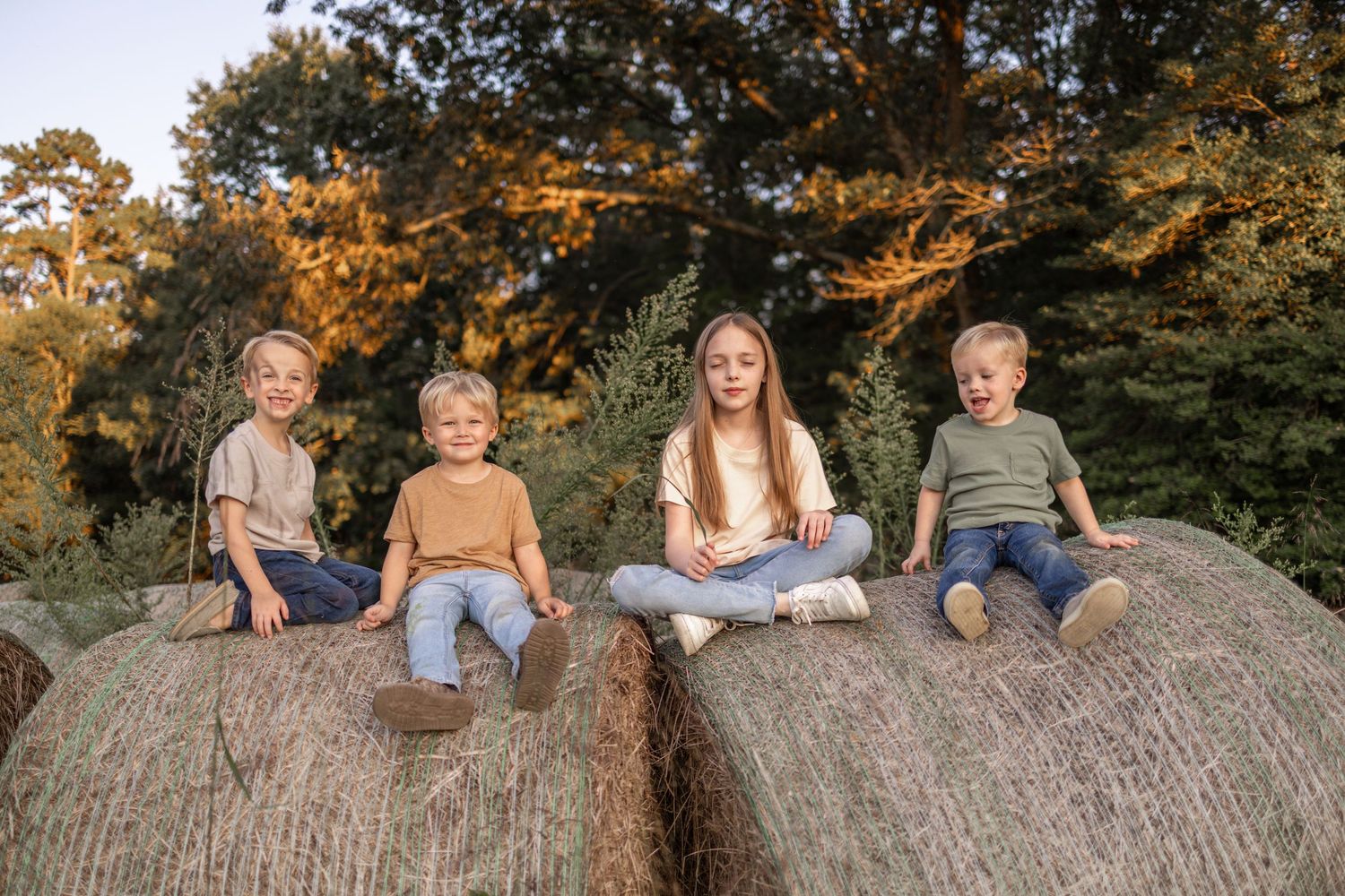 Children sit together on large round hay bales during golden sunset hour in autumn.