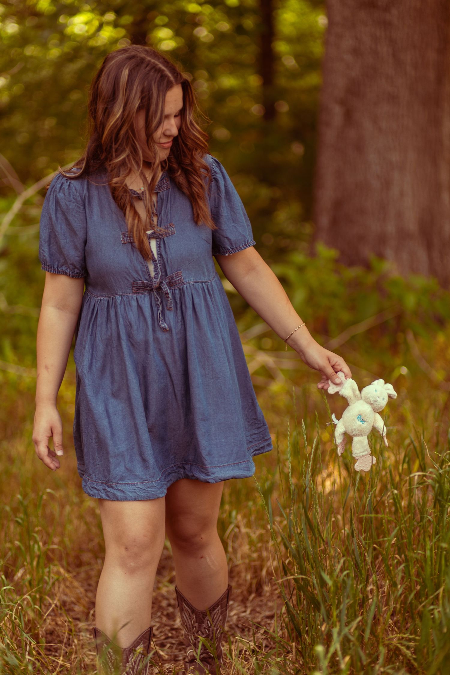 Young woman in blue denim dress and cowboy boots holding a small toy unicorn in a lush forest setting.