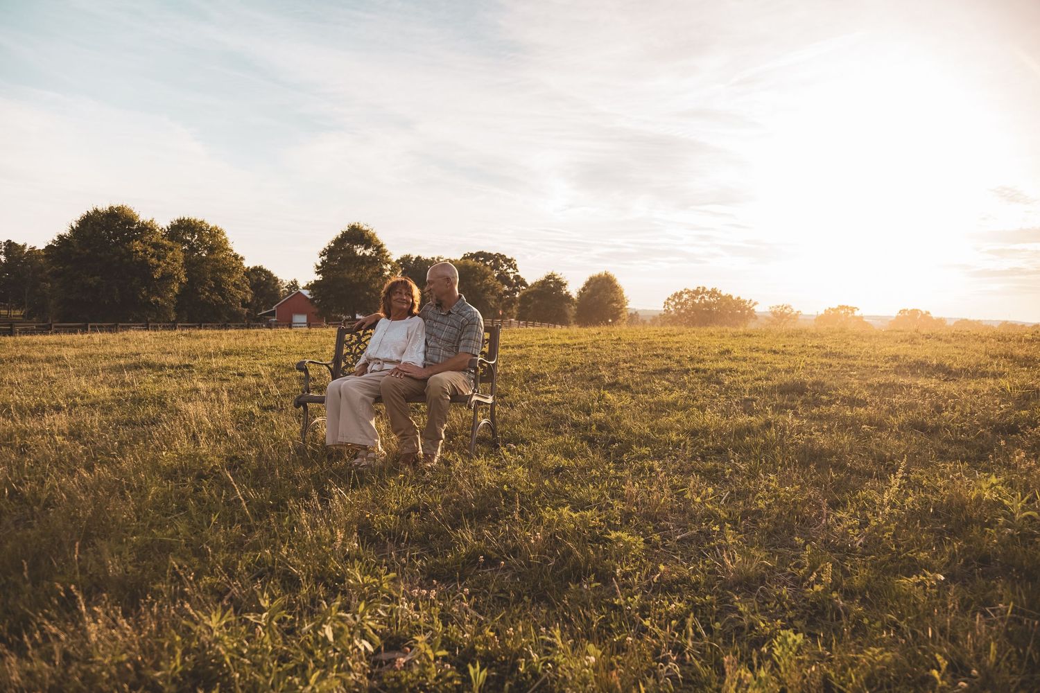 Elderly couple sitting on a bench in a sunlit green meadow surrounded by trees at golden hour.