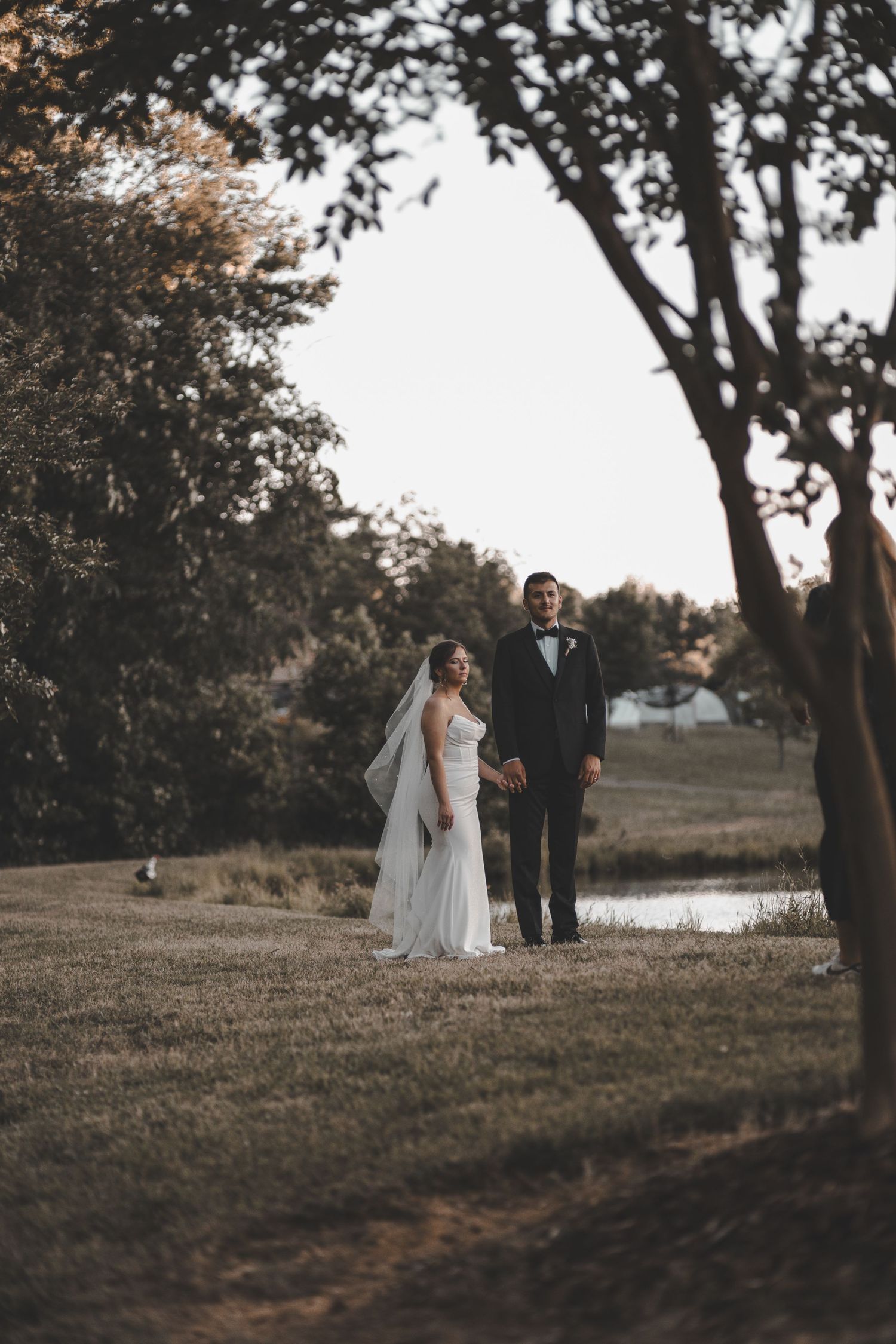 A wedding couple stands together on a scenic hillside overlooking a pond at dusk.