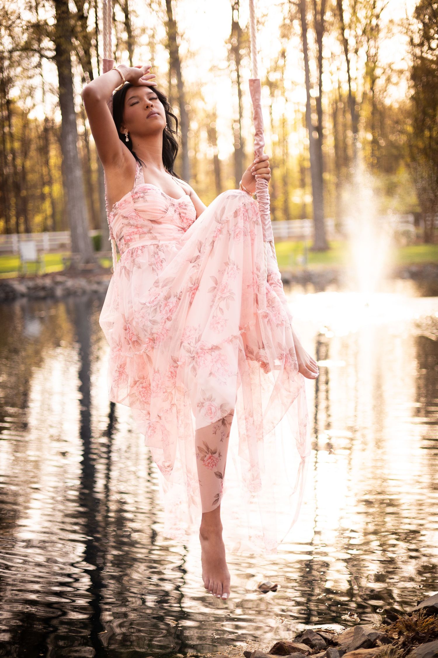 Woman in floral pink gown standing in water, posing gracefully among trees with golden light in background.