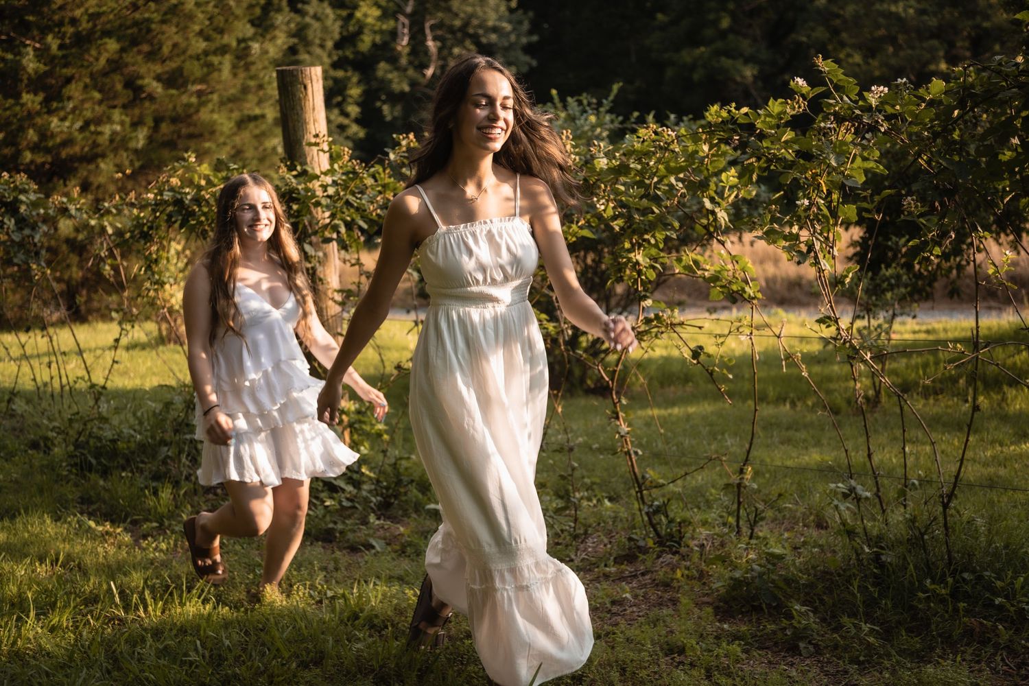 Two people in flowing white dresses run playfully through a sunlit garden on a summer day.