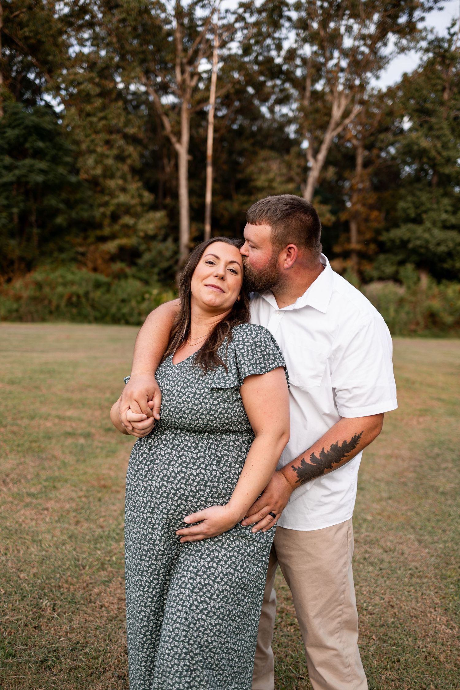 A couple shares a romantic moment outdoors in a grassy field with trees in the background.