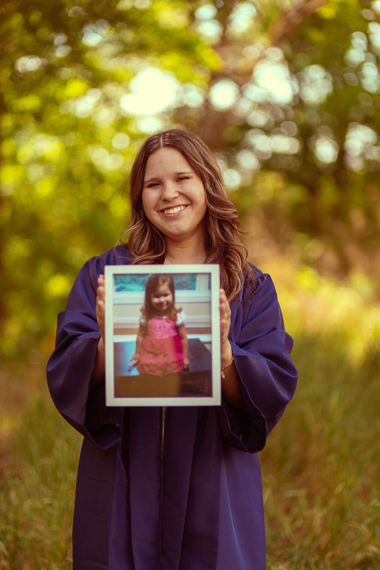 Smiling graduate in blue gown holds childhood photo outdoors among green trees.