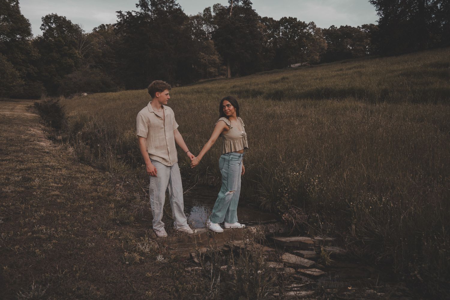 A couple holds hands while standing on a rustic log path in a field at sunset.
