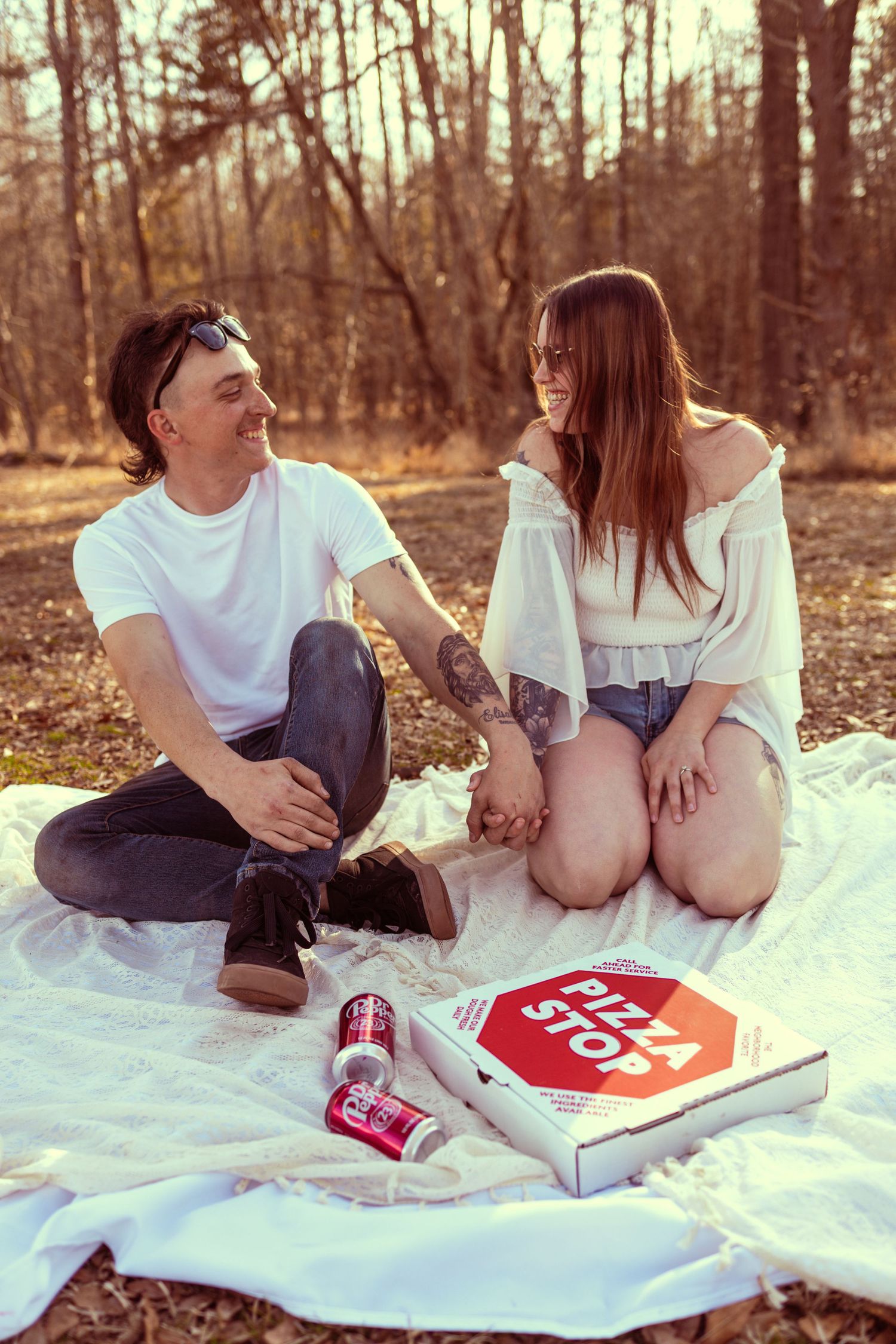 Couple enjoying a romantic outdoor picnic on a white blanket with Pizza Stop box and drinks in a forest setting.