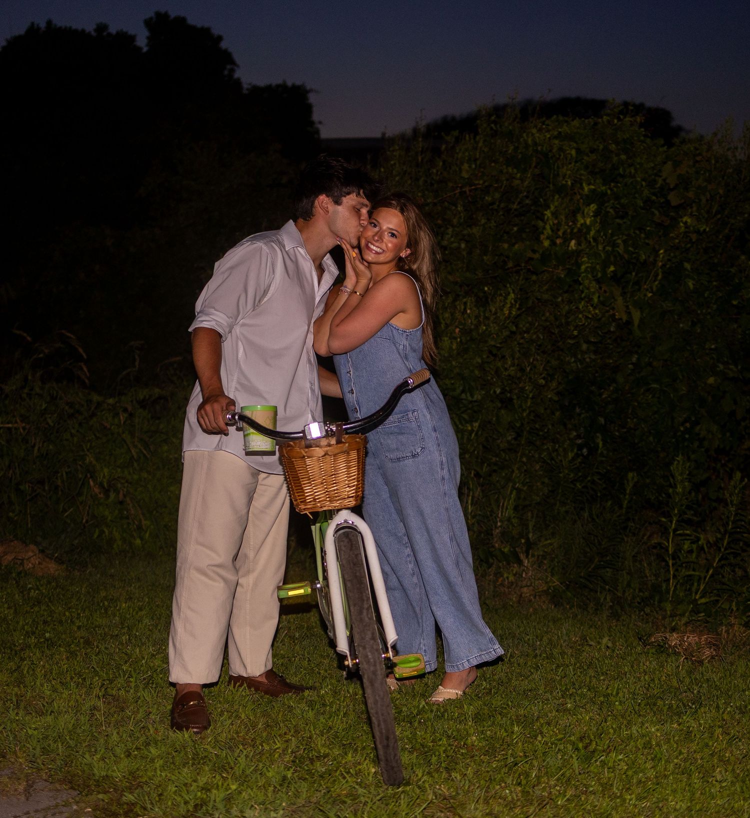 A couple shares a romantic moment at dusk beside a bicycle with a wicker basket in a lush green setting.