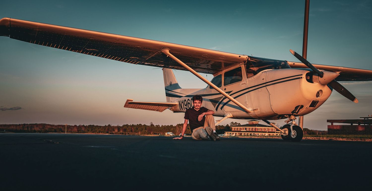 Person crouching beside a white Cessna small plane on a runway at sunset with a colorful sky backdrop.