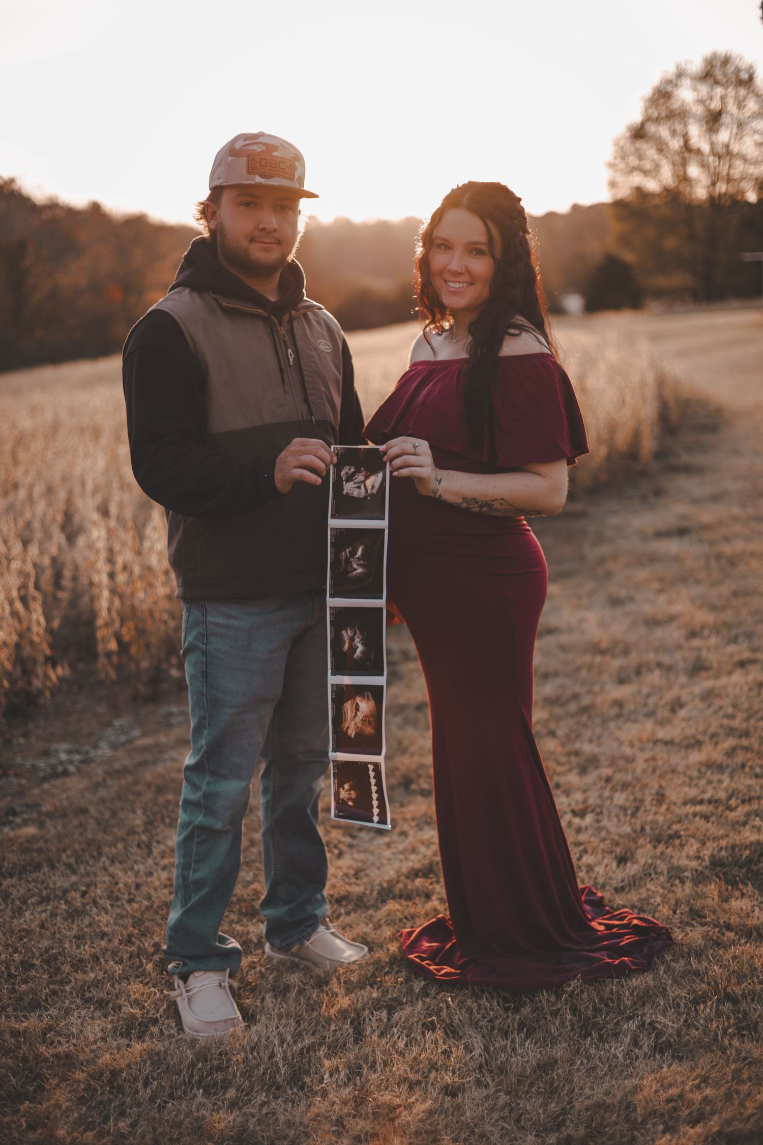 A couple stands in a golden field at sunset holding ultrasound photos during their pregnancy announcement photoshoot.