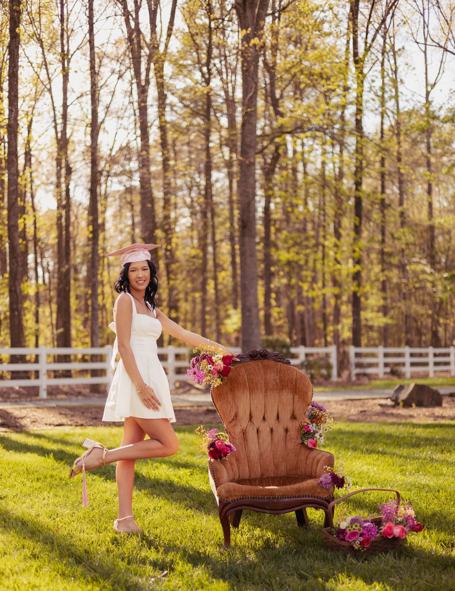 Woman in white dress and pink hat poses beside a vintage velvet chair adorned with pink flowers on a sunny green lawn.