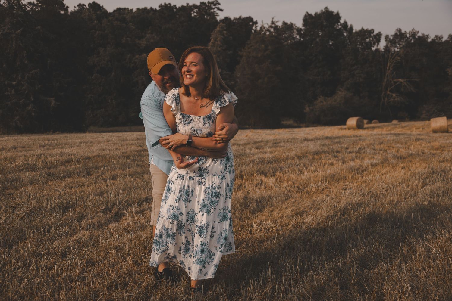 A couple shares a tender moment while standing in a rustic field with hay bales.