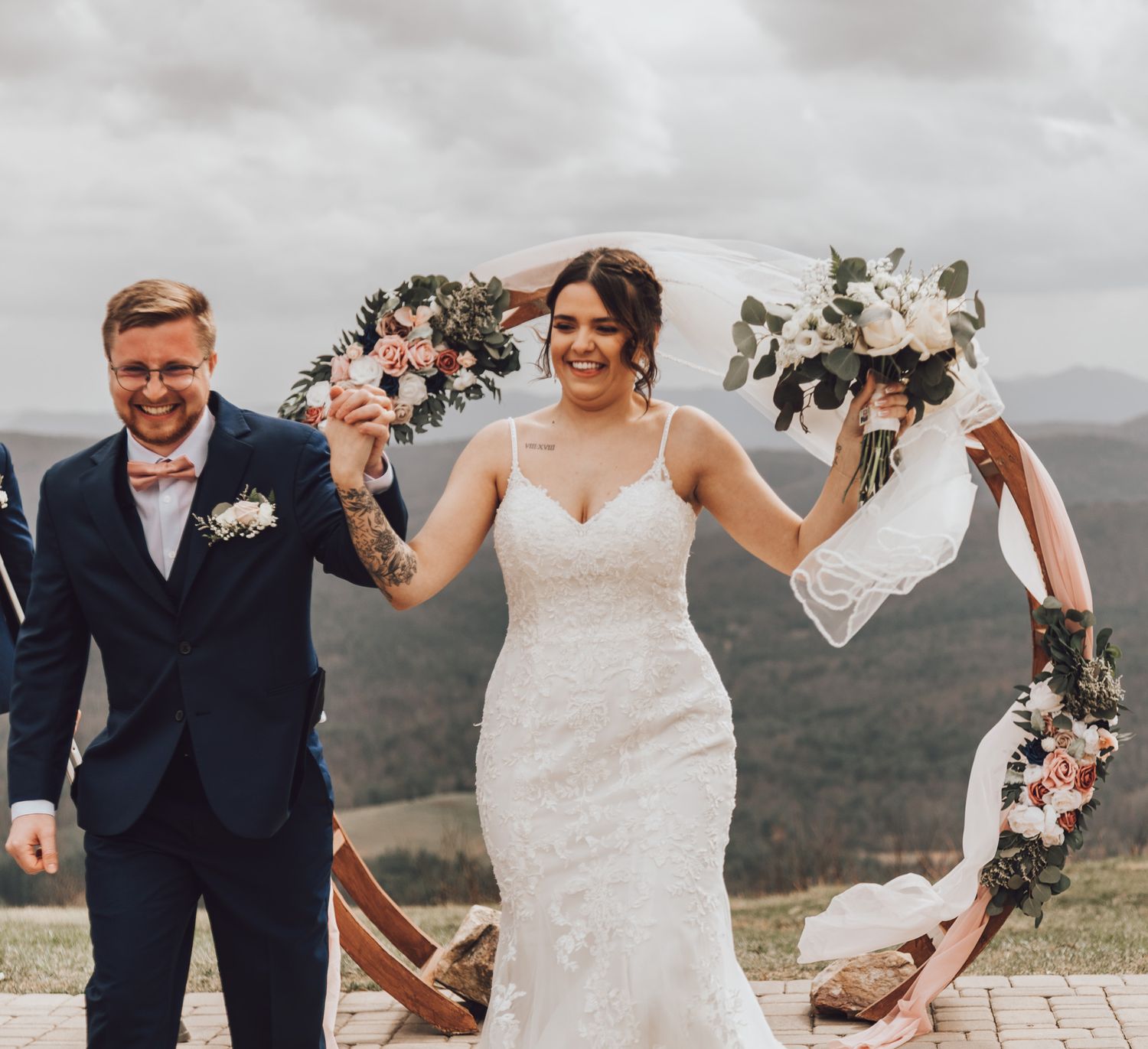 Wedding couple celebrates joyfully on a hillside with white and pink floral bouquets against dramatic cloudy skies.