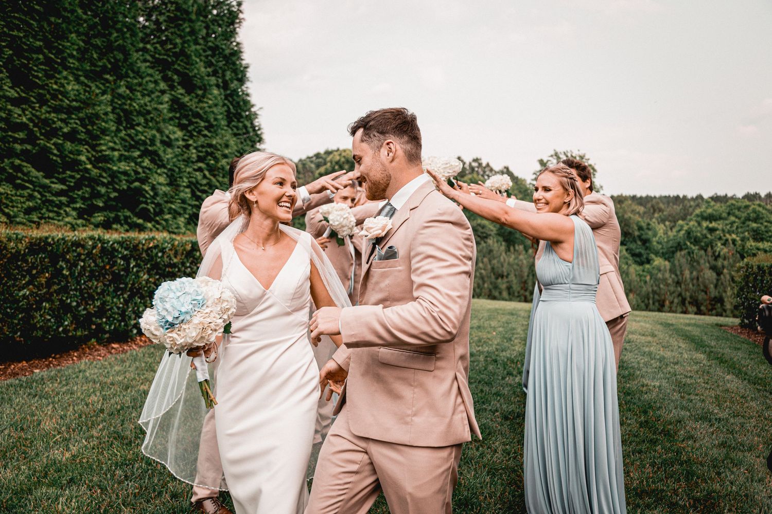Wedding party in tan suits and blue dresses posing playfully on garden grounds.
