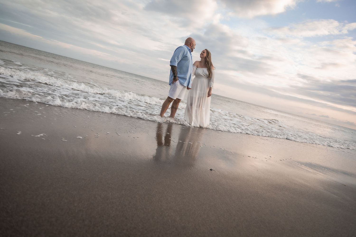 A couple walks together along a misty shoreline as waves crash on the beach at sunset.