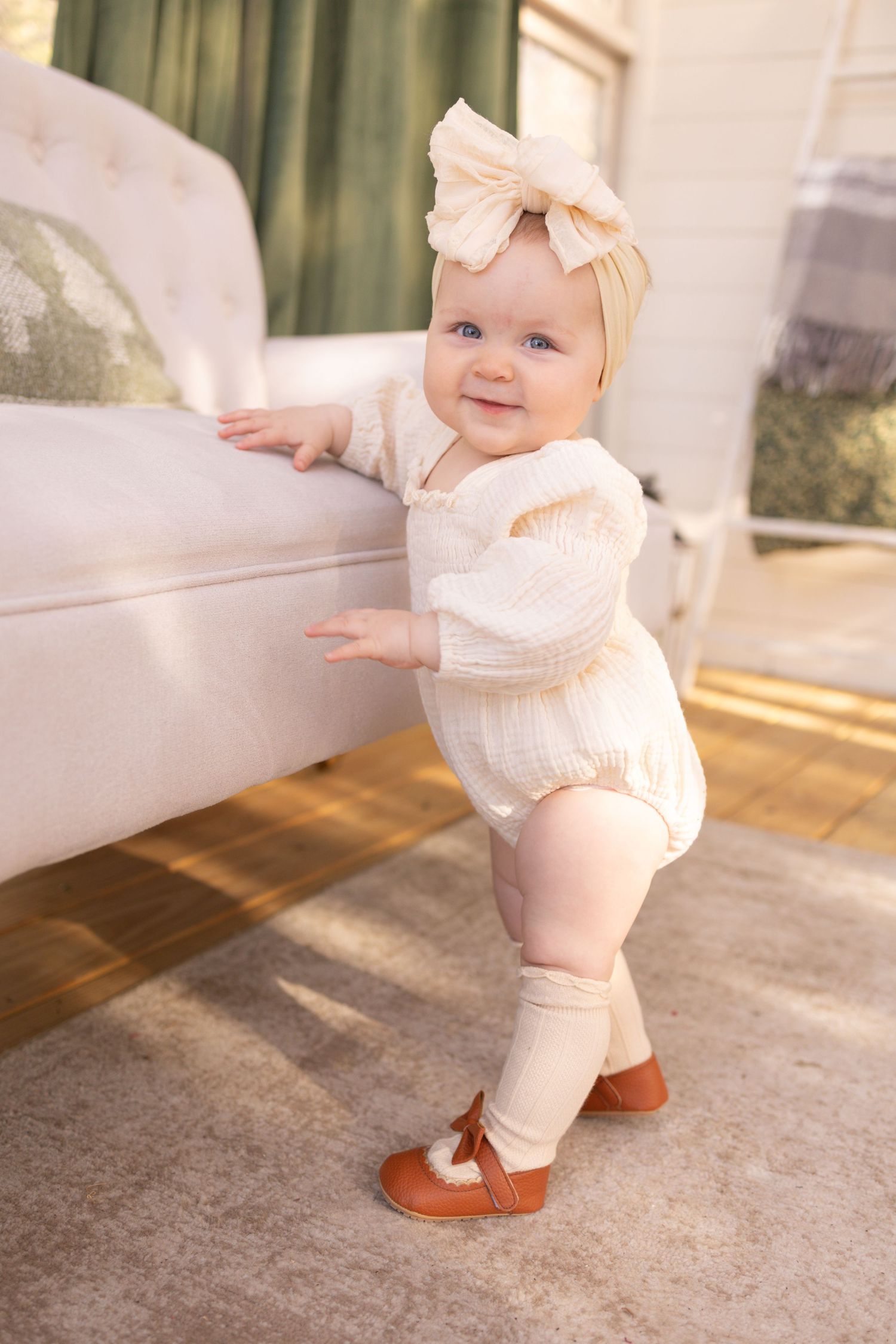 A baby wearing a cream-colored outfit and bow crawls and stands on a carpeted floor in a progression of movements.