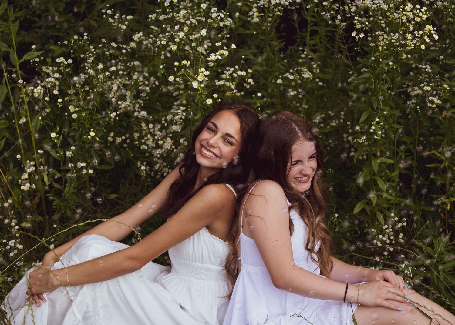 Two friends in white dresses sit back-to-back amid blooming flowers and greenery in a garden setting.