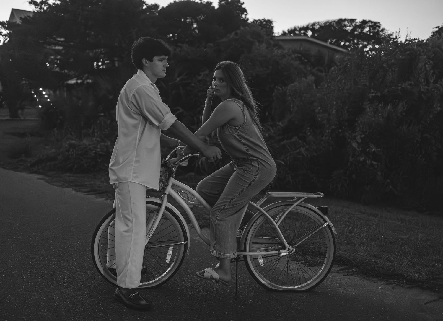 Black and white photo of a young couple sharing a bicycle on a quiet road at dusk near trees.