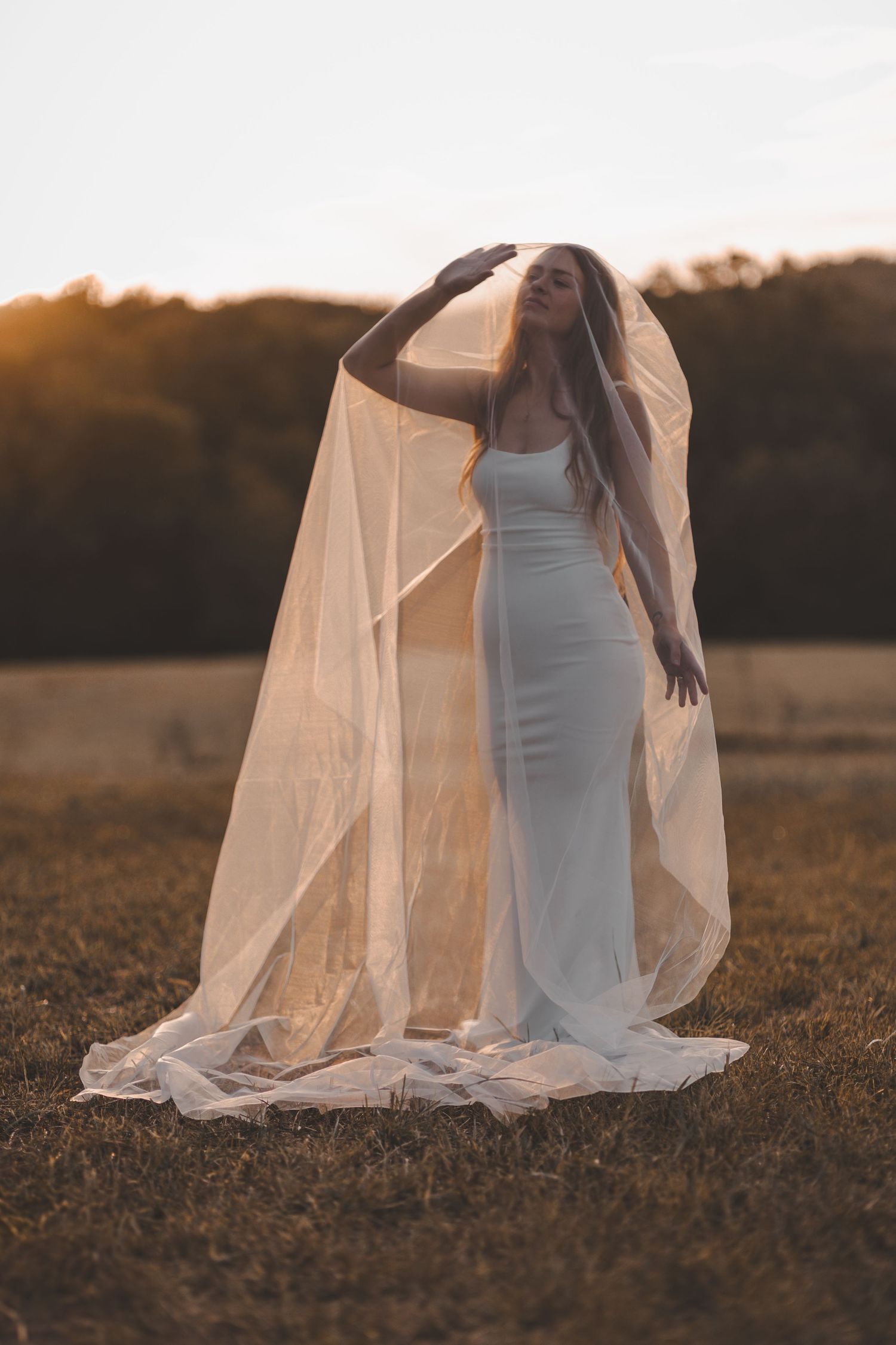 A figure in a white dress and flowing veil poses gracefully in a golden field at sunset.