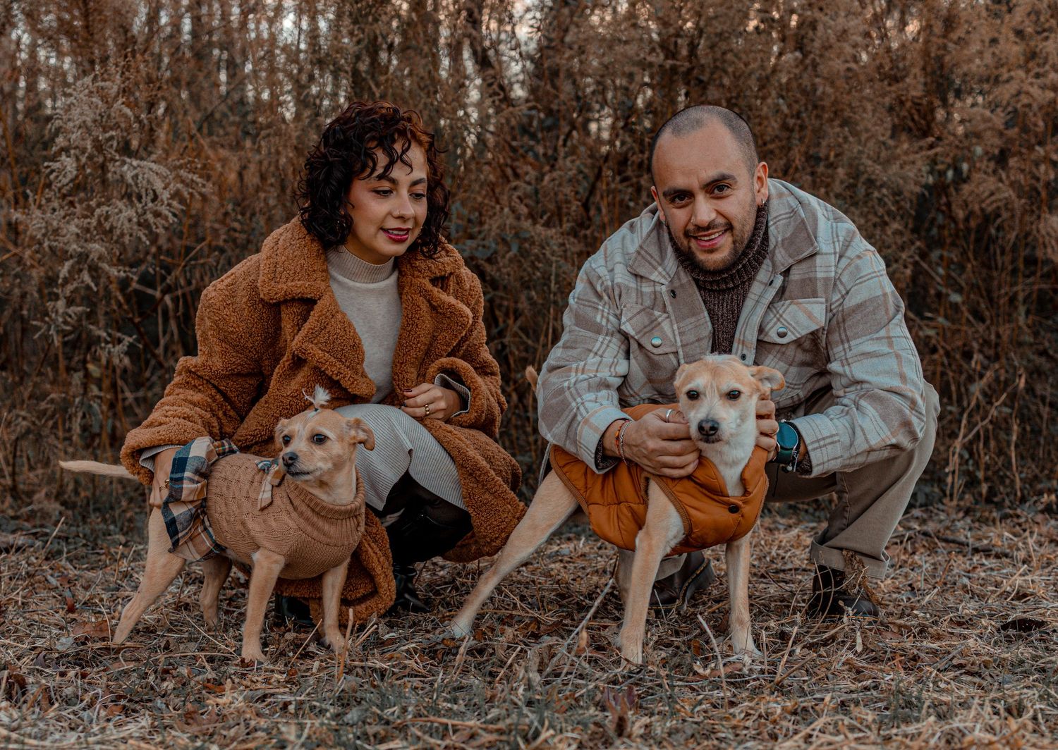 A couple in autumn clothing kneels outdoors with their two tan rescue dogs wearing orange bandanas.