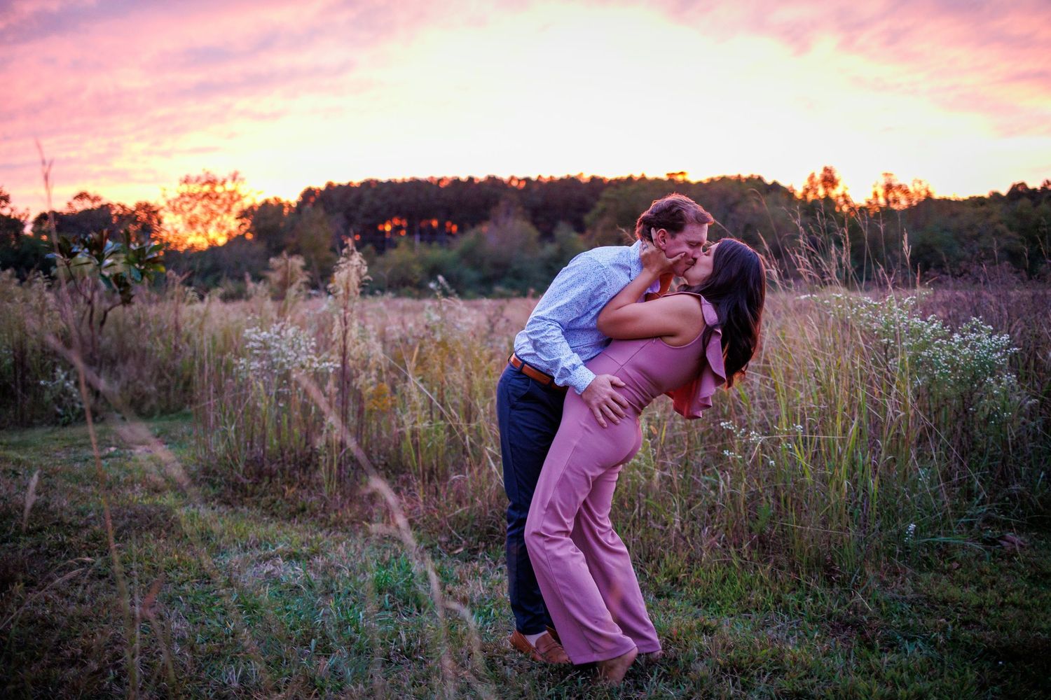 A romantic couple embraces in a field during a stunning pink and orange sunset.