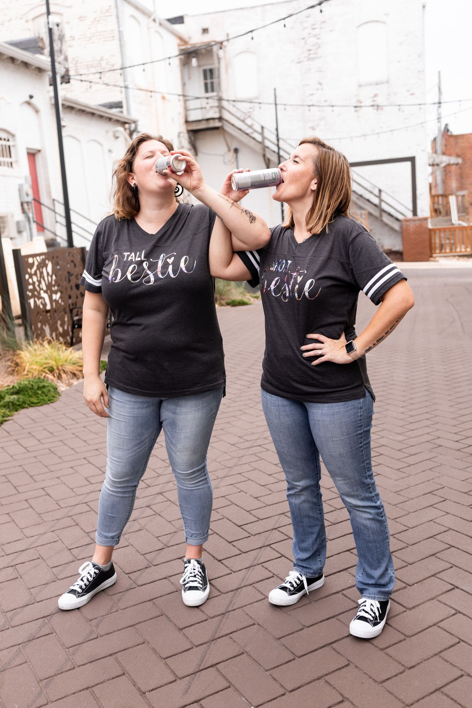 Two people wearing matching black t-shirts and jeans strike playful poses on a brick pavement in an urban setting.