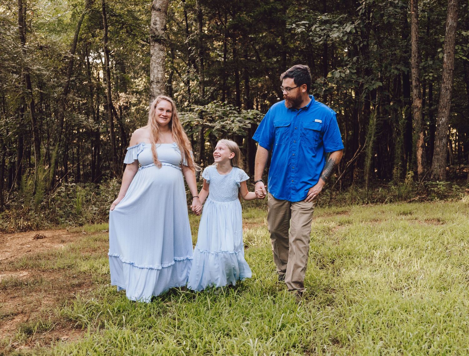 A family shares tender moments on a grassy slope in a wooded area, wearing coordinated light blue and navy outfits.