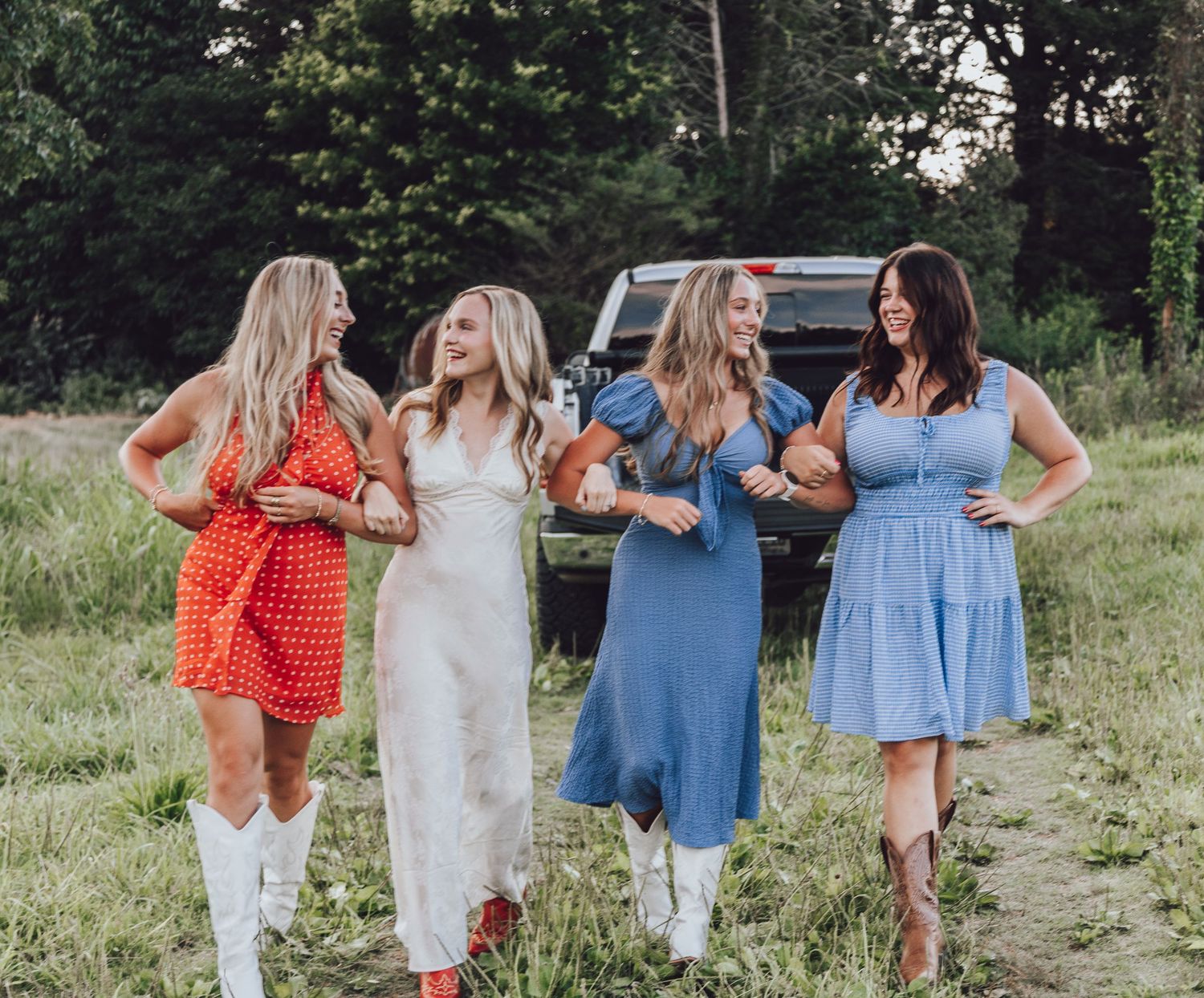 Four friends in summer dresses and white boots stand together laughing in a grassy field with a truck in background.