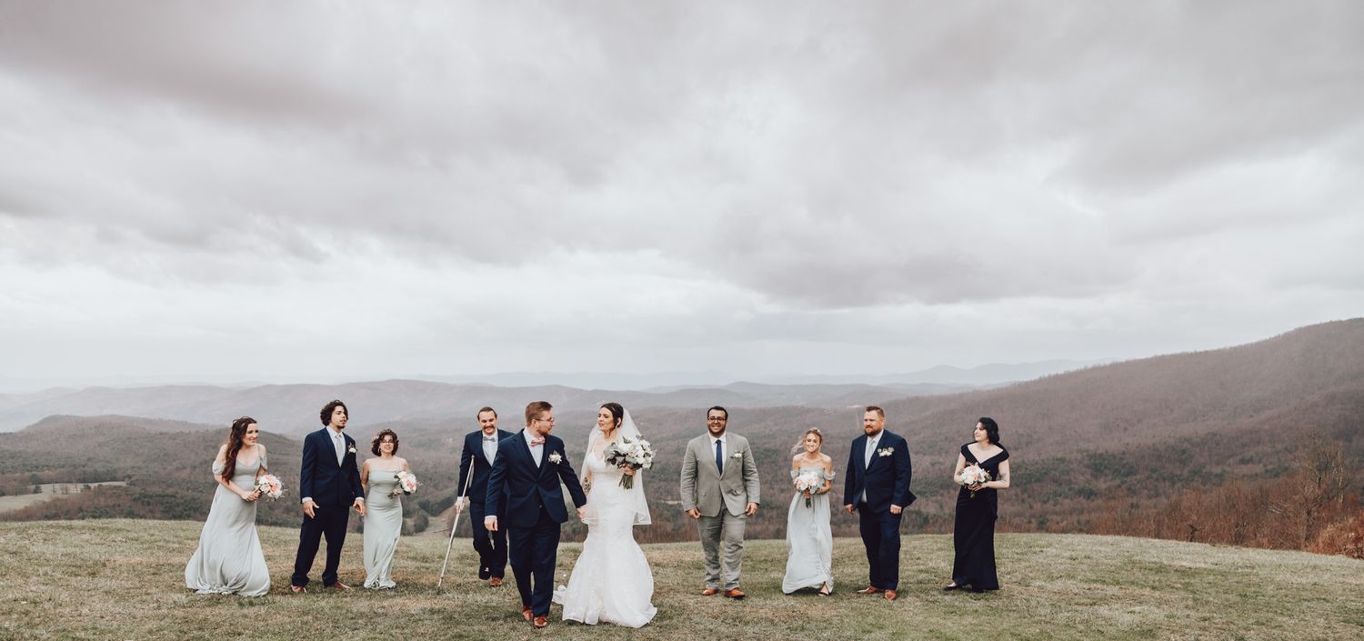 Wedding party poses for group photo on scenic mountain overlook with cloudy dramatic sky.