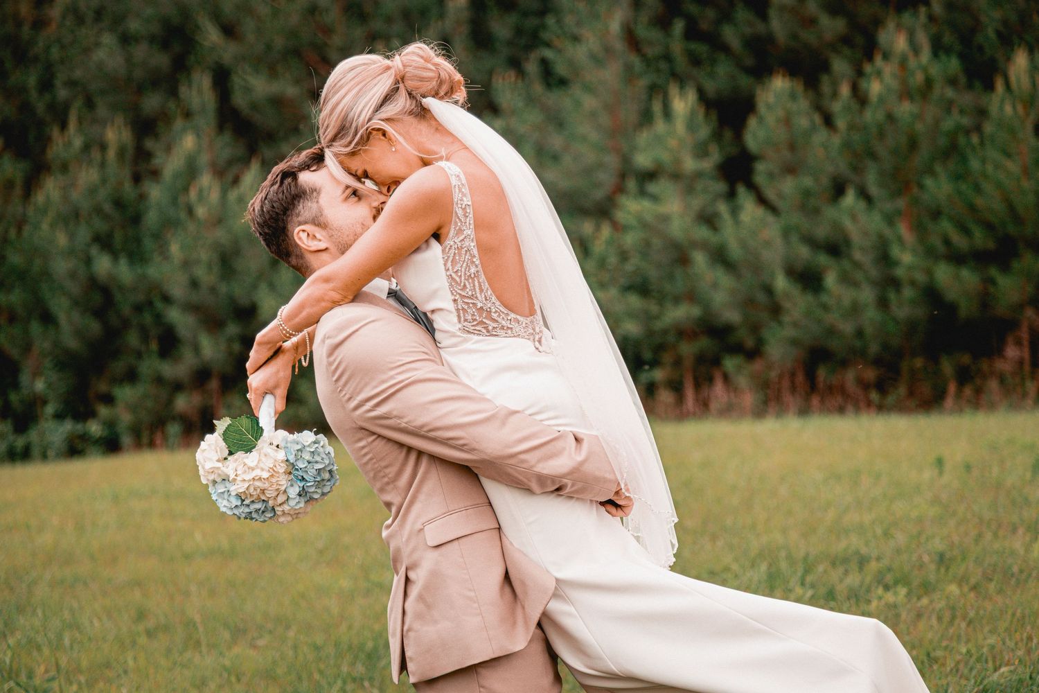 A romantic wedding moment captured outdoors as a couple embraces in a joyful lift pose against a natural backdrop.