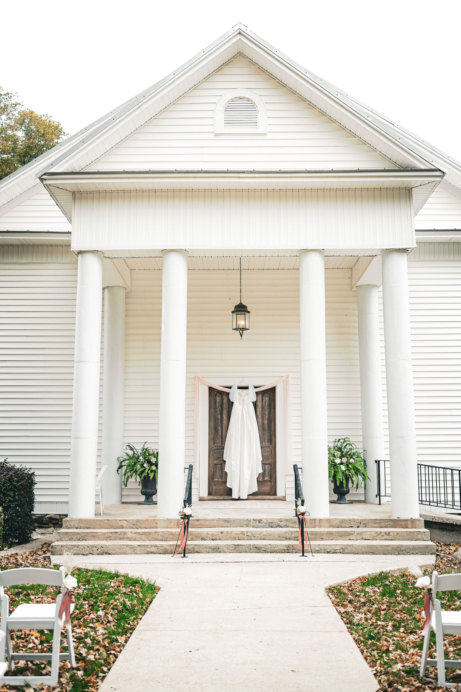 White colonial wedding venue with hanging bridal gown, white columns, and chairs lining the pathway on an autumn day.