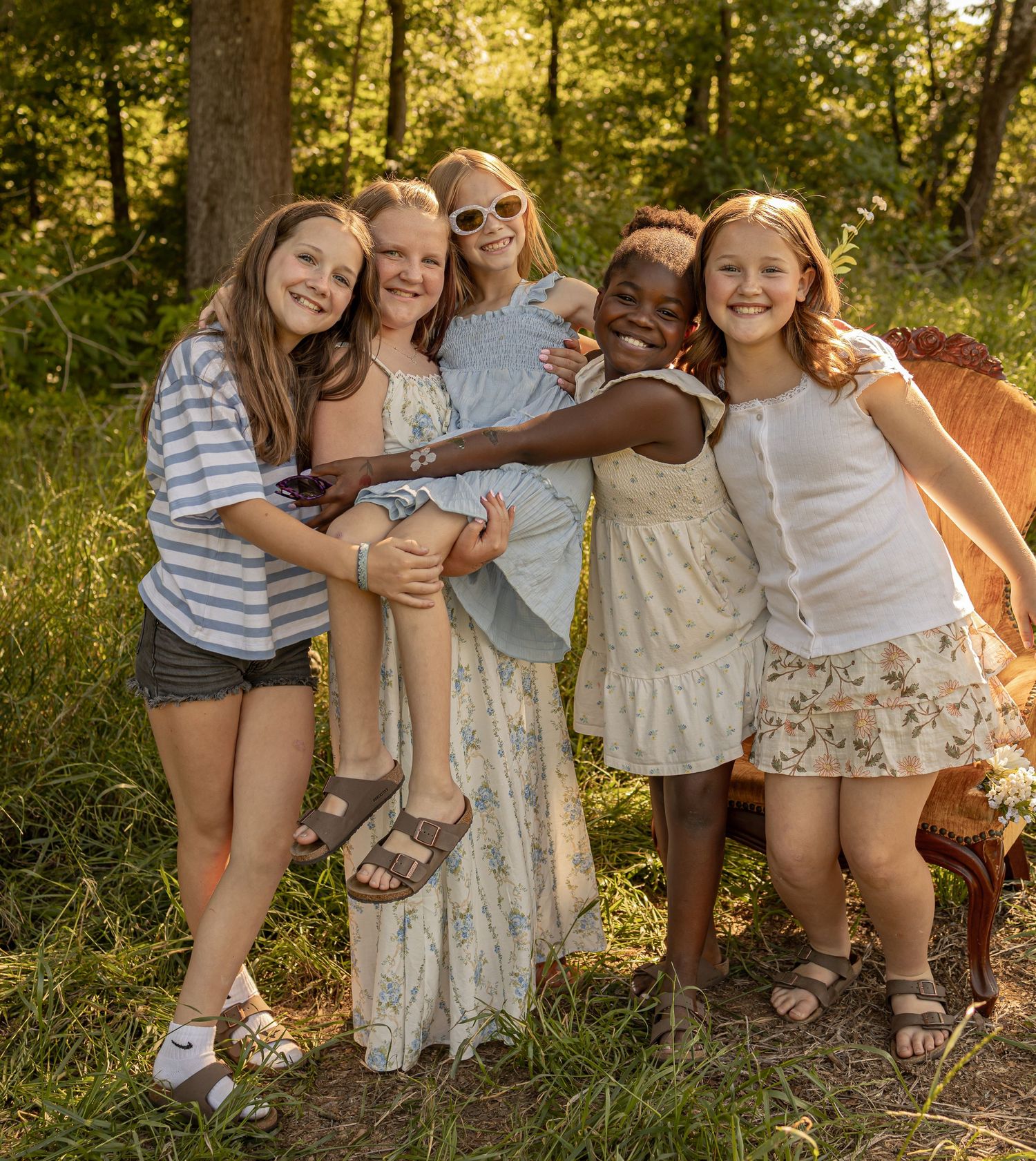 Five young girls hugging and smiling together outdoors in a sunny forest setting.
