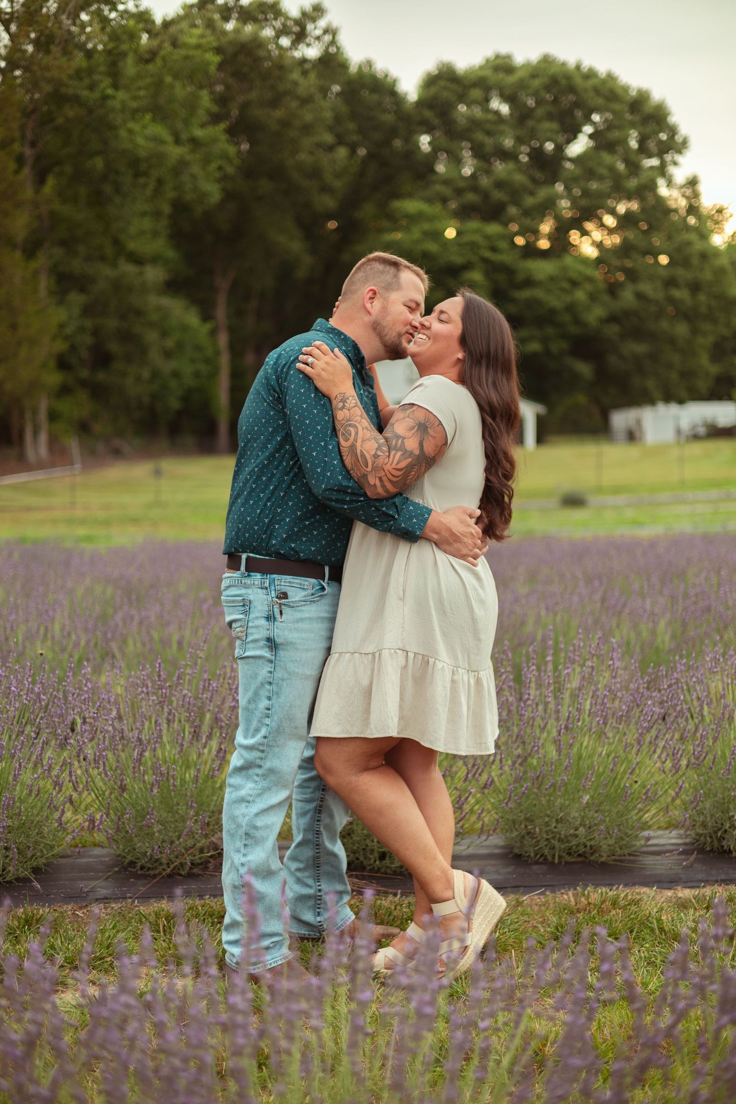 A couple shares a romantic kiss in a blooming lavender field, the man in a teal shirt and the woman in a white dress.