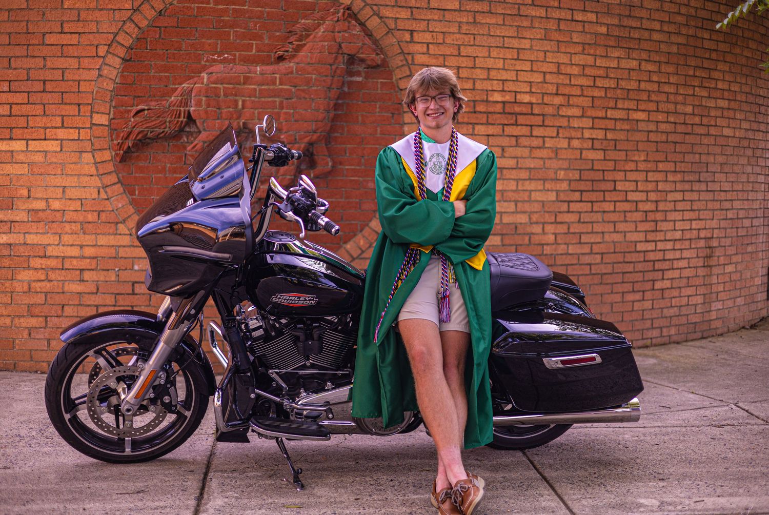 Graduate in green gown poses beside a Harley-Davidson motorcycle against a brick wall backdrop.