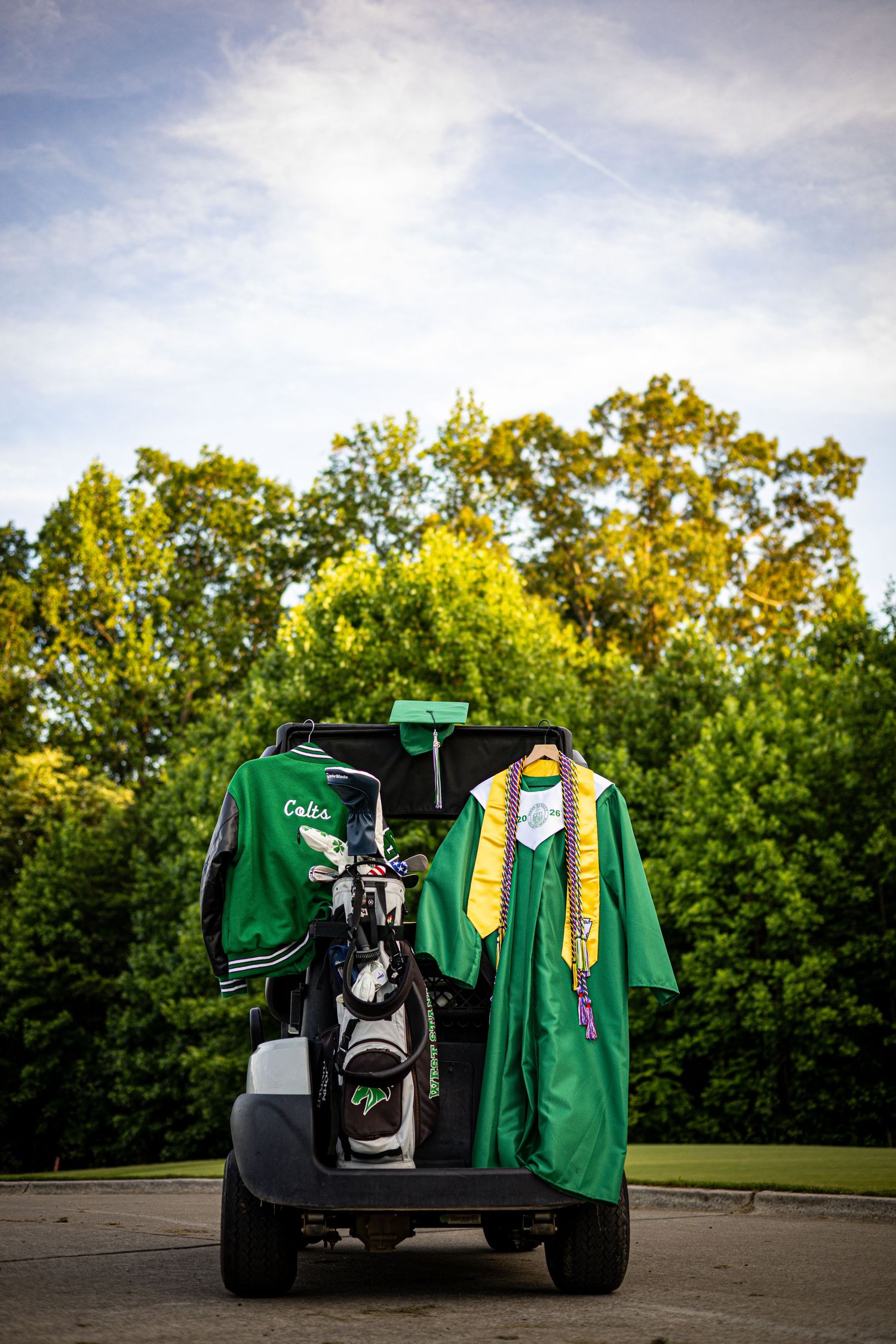 Green graduation gown and jacket hanging on golf cart with trees in background.
