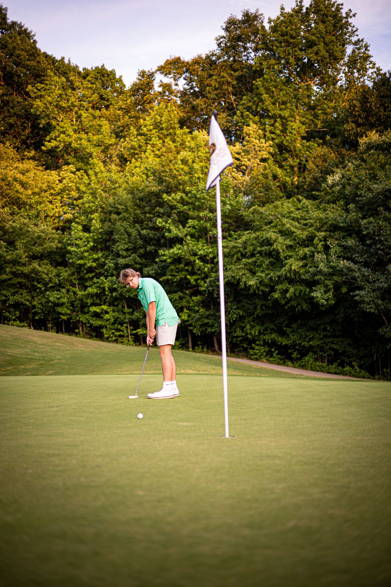 Golfer in green shirt putting on a lush golf course green with white flag pin and trees in background.