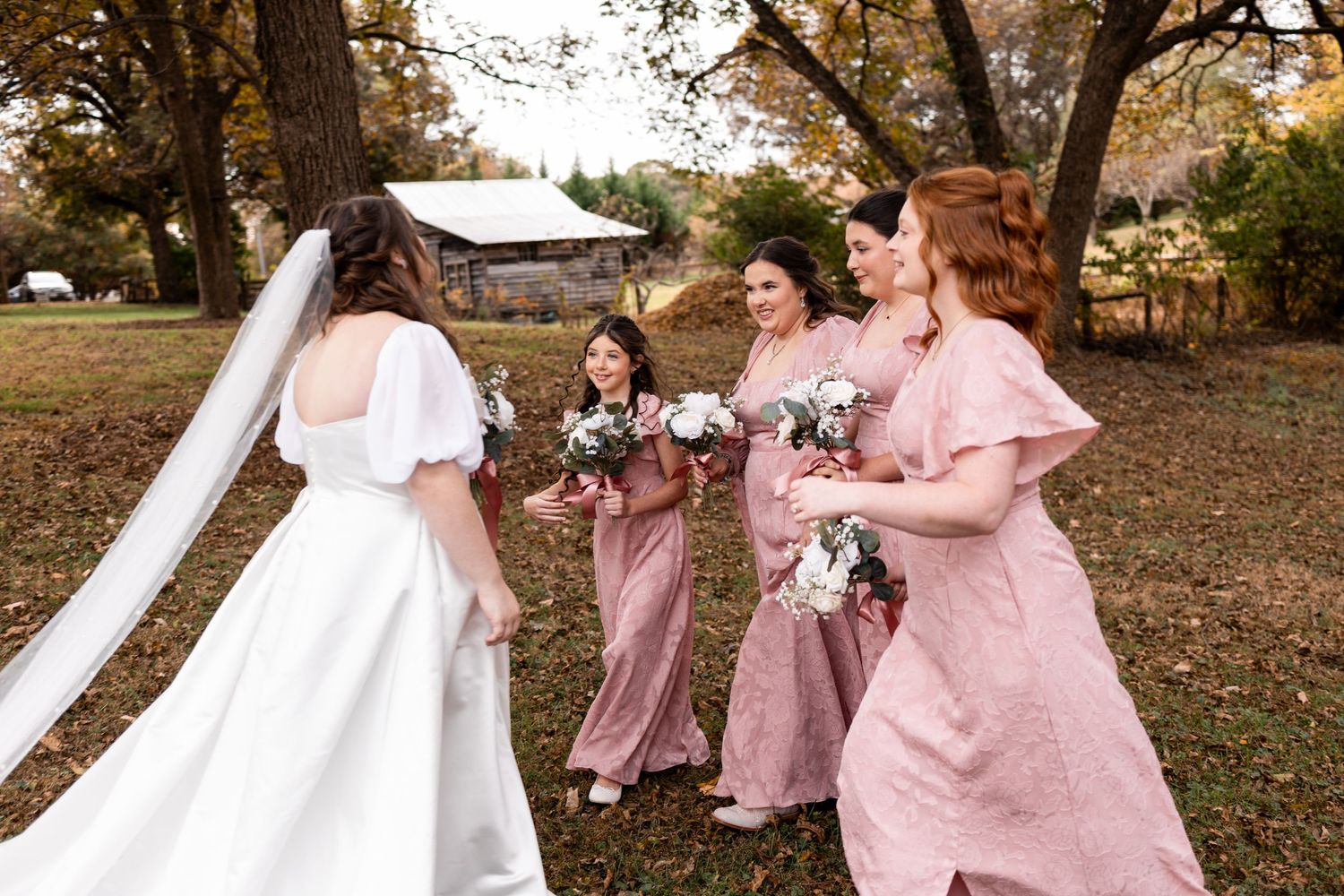 Bride in white gown with veil walks alongside three bridesmaids in pink dresses holding bouquets outdoors.