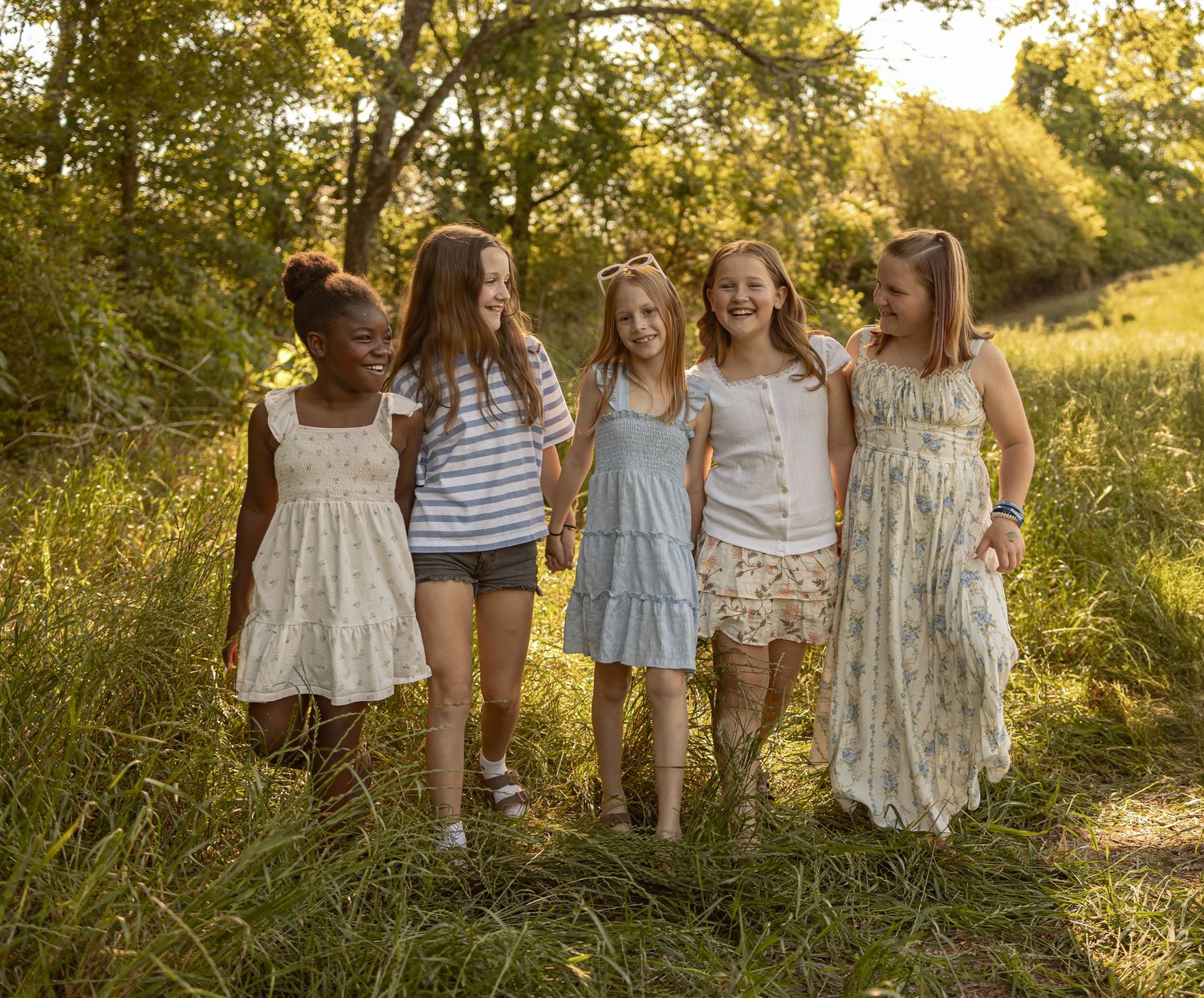 Five girls walking together laughing in a sunny meadow surrounded by lush green trees and tall grass.