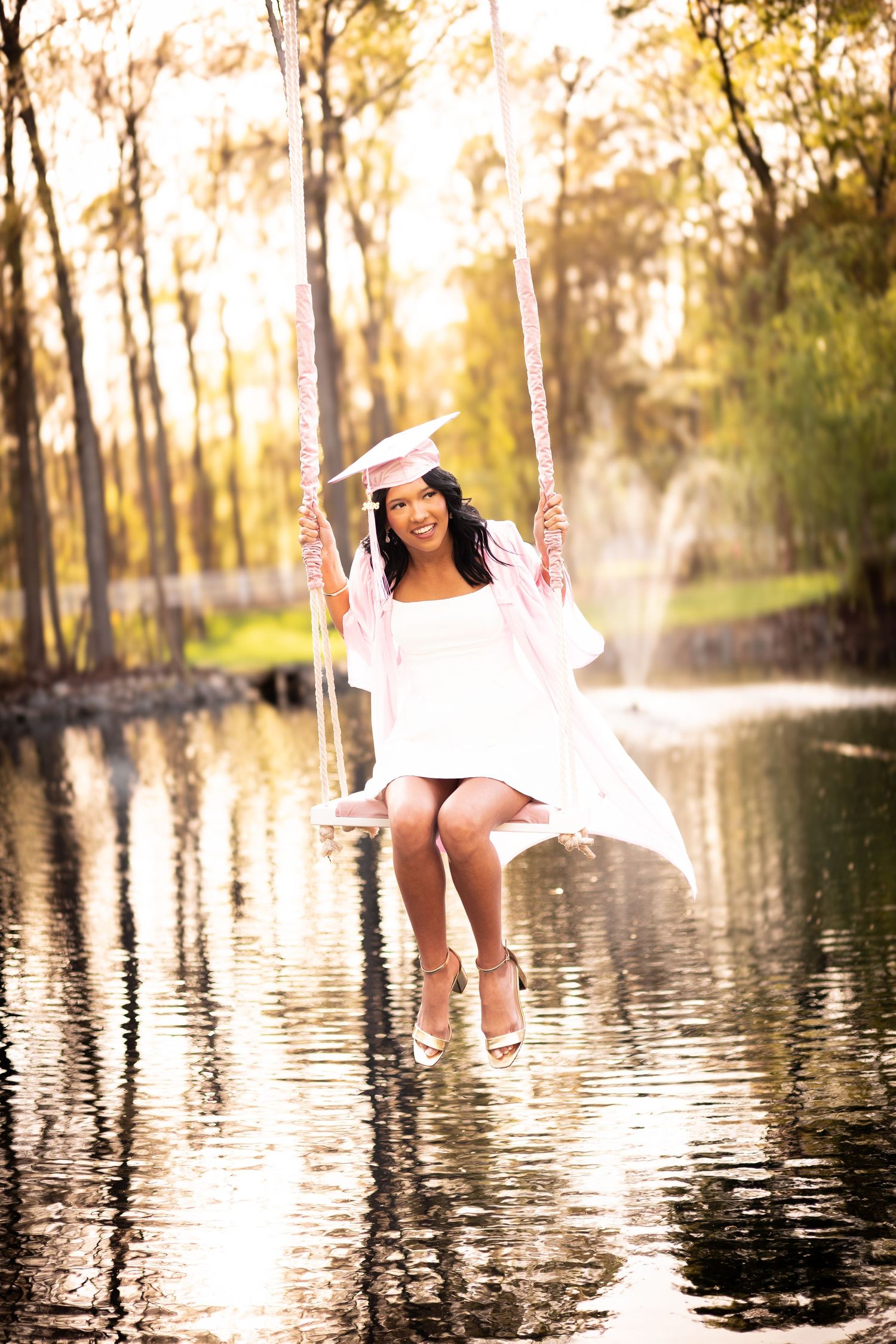 Young woman in white graduation cap and gown smiling on a rope swing over a reflective pond surrounded by autumn trees.