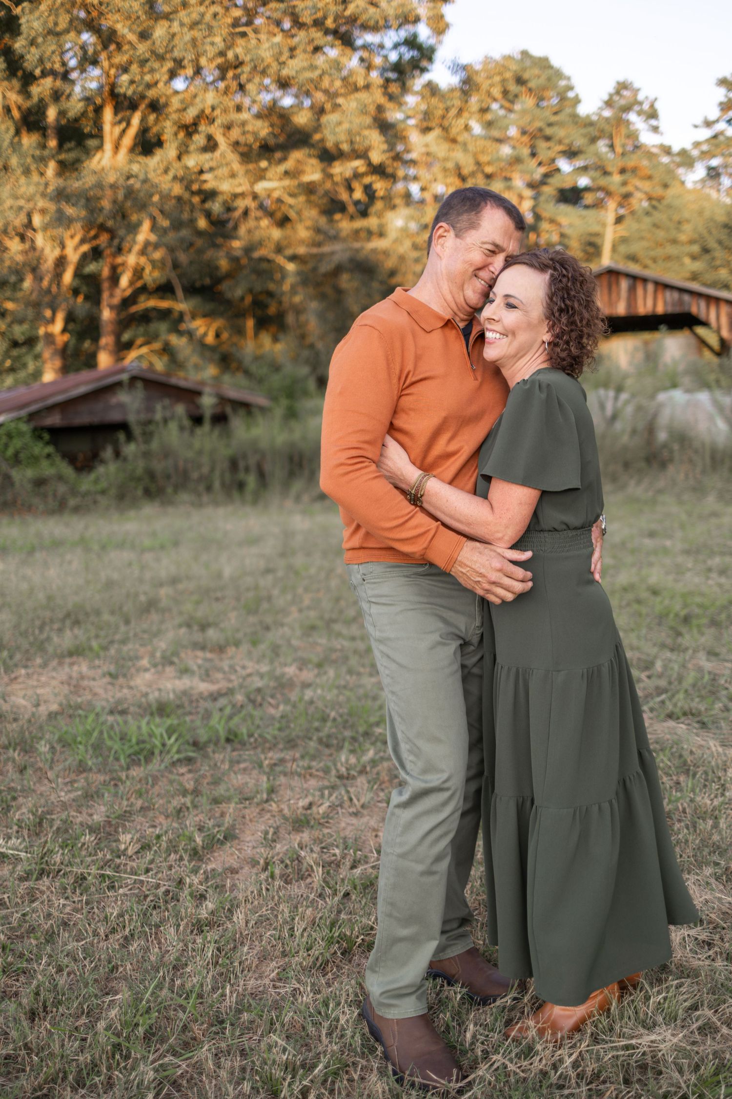A couple embraces and laughs together in a grassy field during golden hour with a rustic barn in the background.