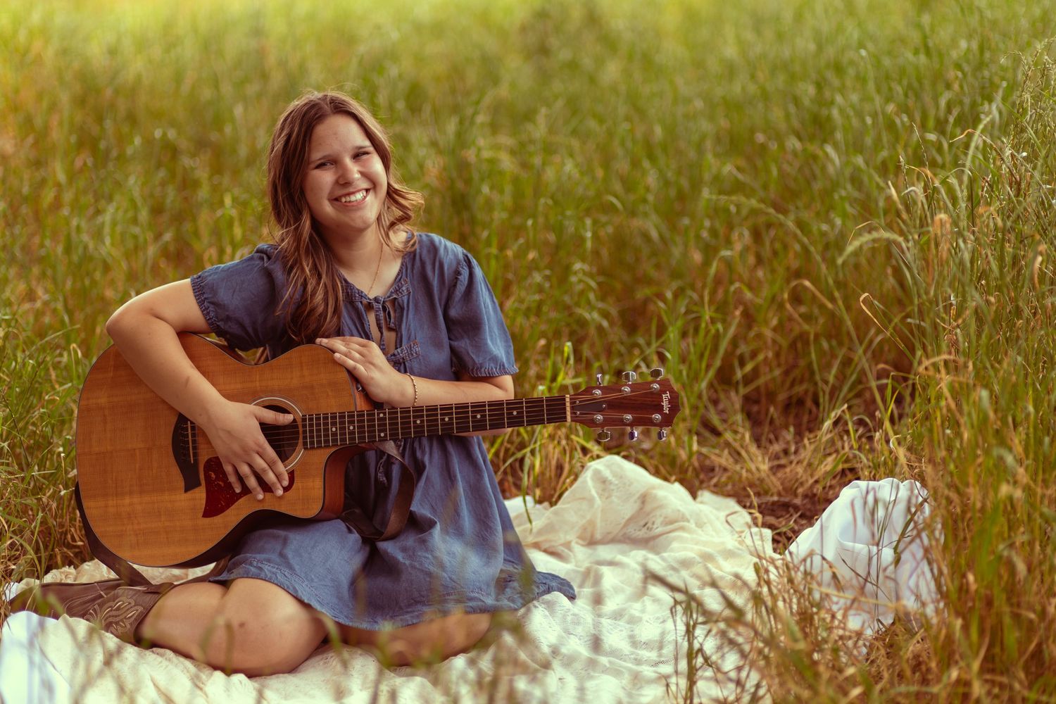 Woman in blue dress smiling while playing acoustic guitar outdoors in a lush green field on a blanket.