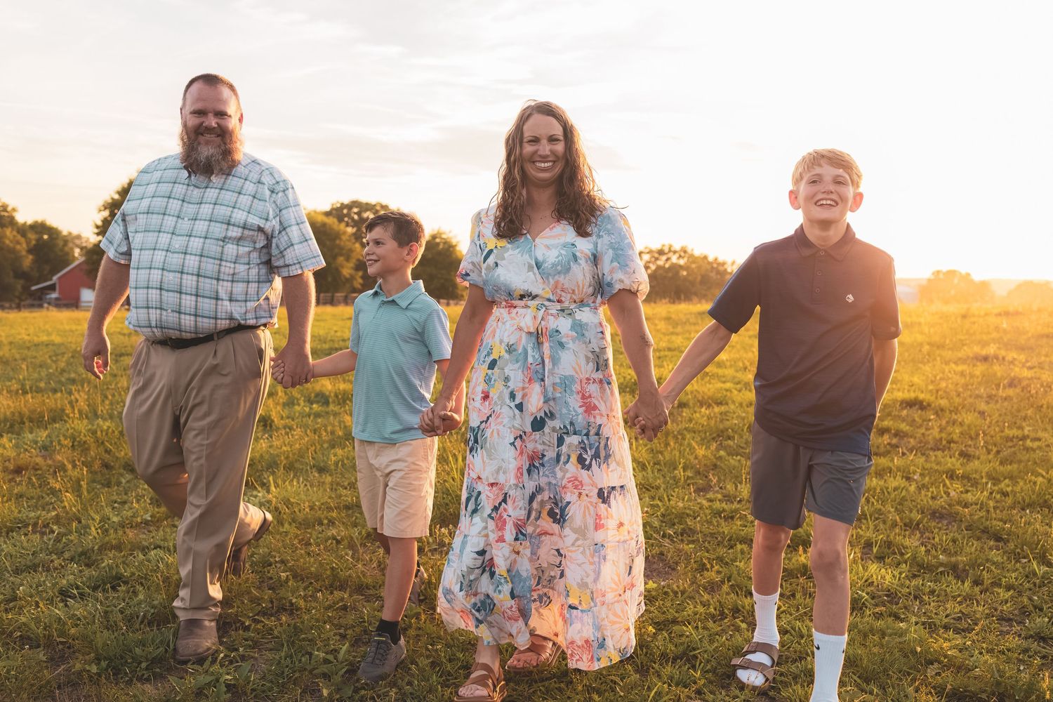Family of four walking hand-in-hand through a sunny green field at golden hour sunset.