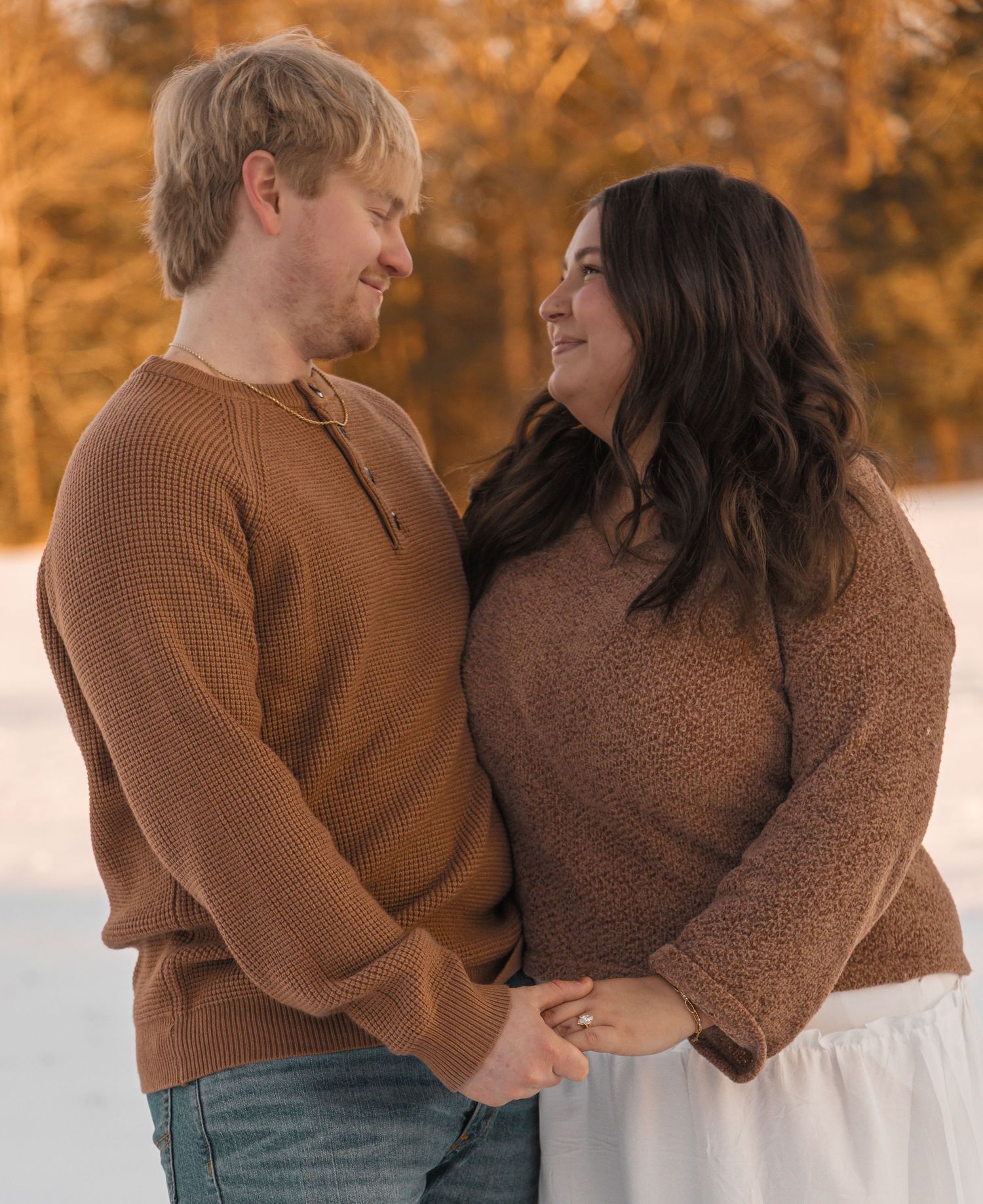 Couple holding hands smiling at each other outdoors in winter, wearing matching brown sweaters with snowy background.