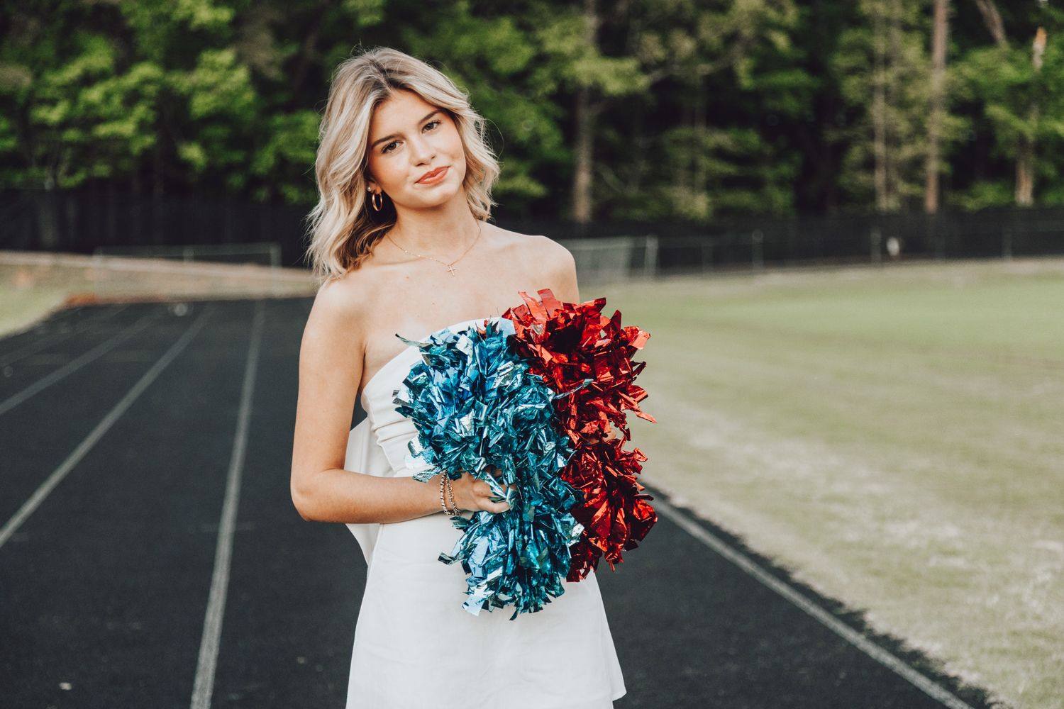 Cheerleader holding blue and red pom poms in white dress on an athletic field.