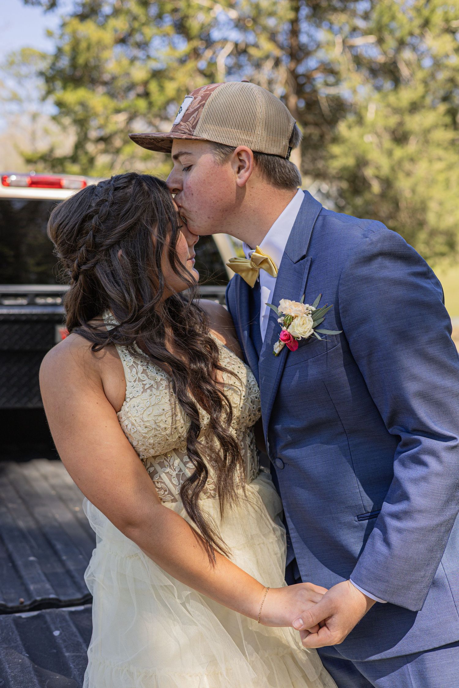 Bride and groom sharing a kiss outdoors, groom wearing blue suit with boutonniere and camo hat, bride in white lace gown.