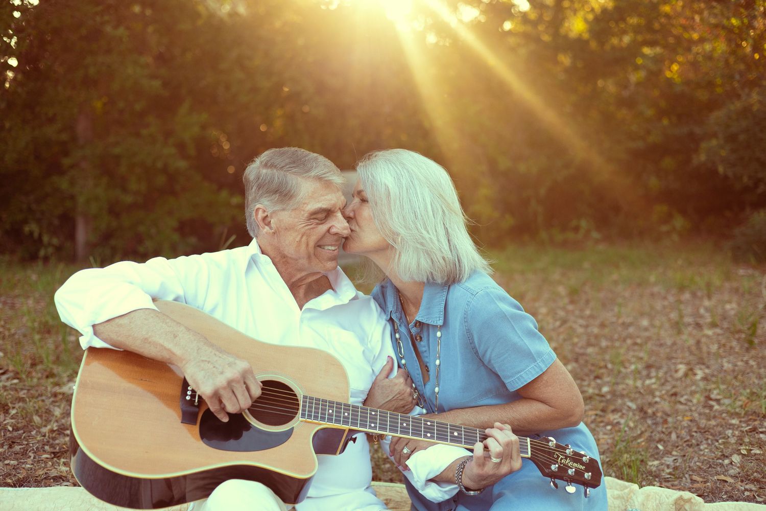 Elderly couple sharing a romantic moment outdoors, man playing acoustic guitar while woman kisses his cheek in golden sunlight.