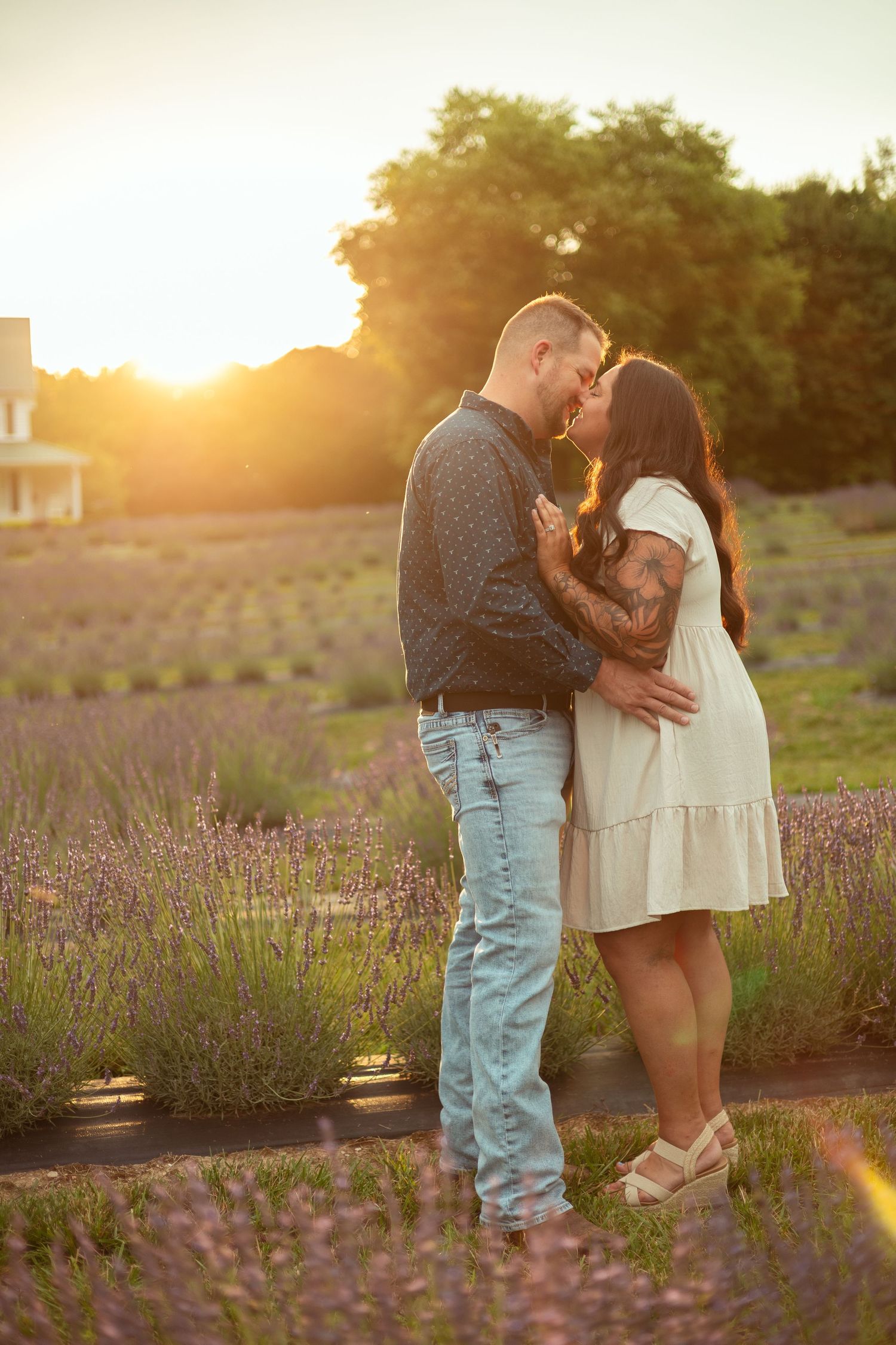 Couple sharing a romantic kiss in a lavender field at golden sunset, woman in white dress, man in jeans.