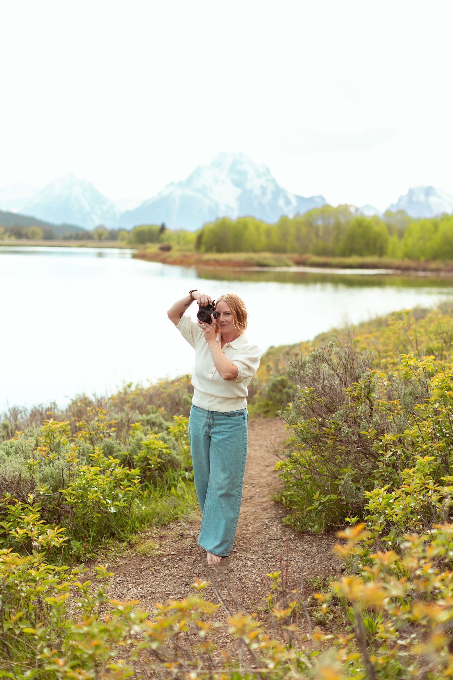 Woman photographer taking photos on a scenic trail near a lake with snow-capped Teton mountains in the background.
