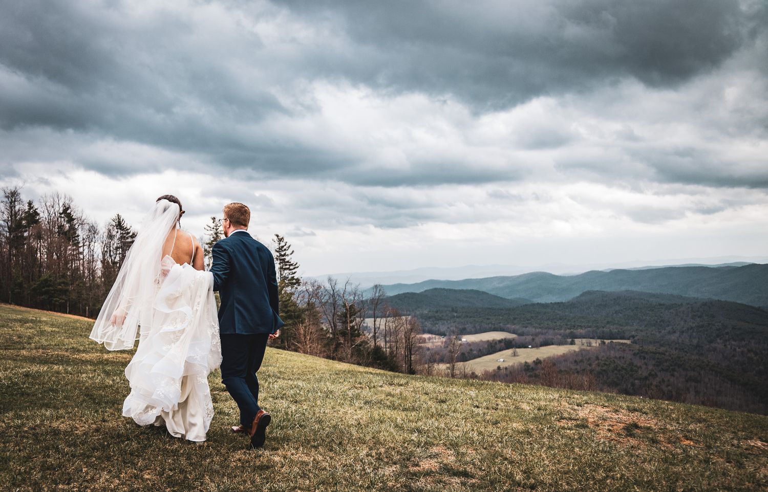 Newlyweds walk together on hilltop meadow with dramatic stormy sky and mountain views in background.