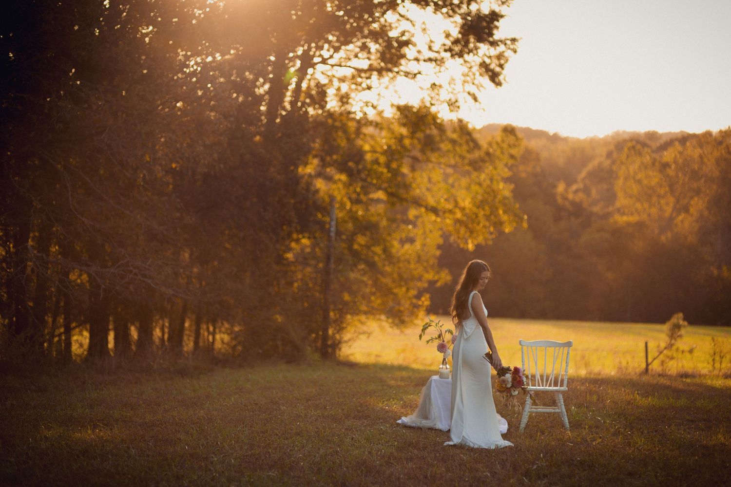 A bride in a white wedding dress sits on a wooden chair in a golden sunlit field during sunset.