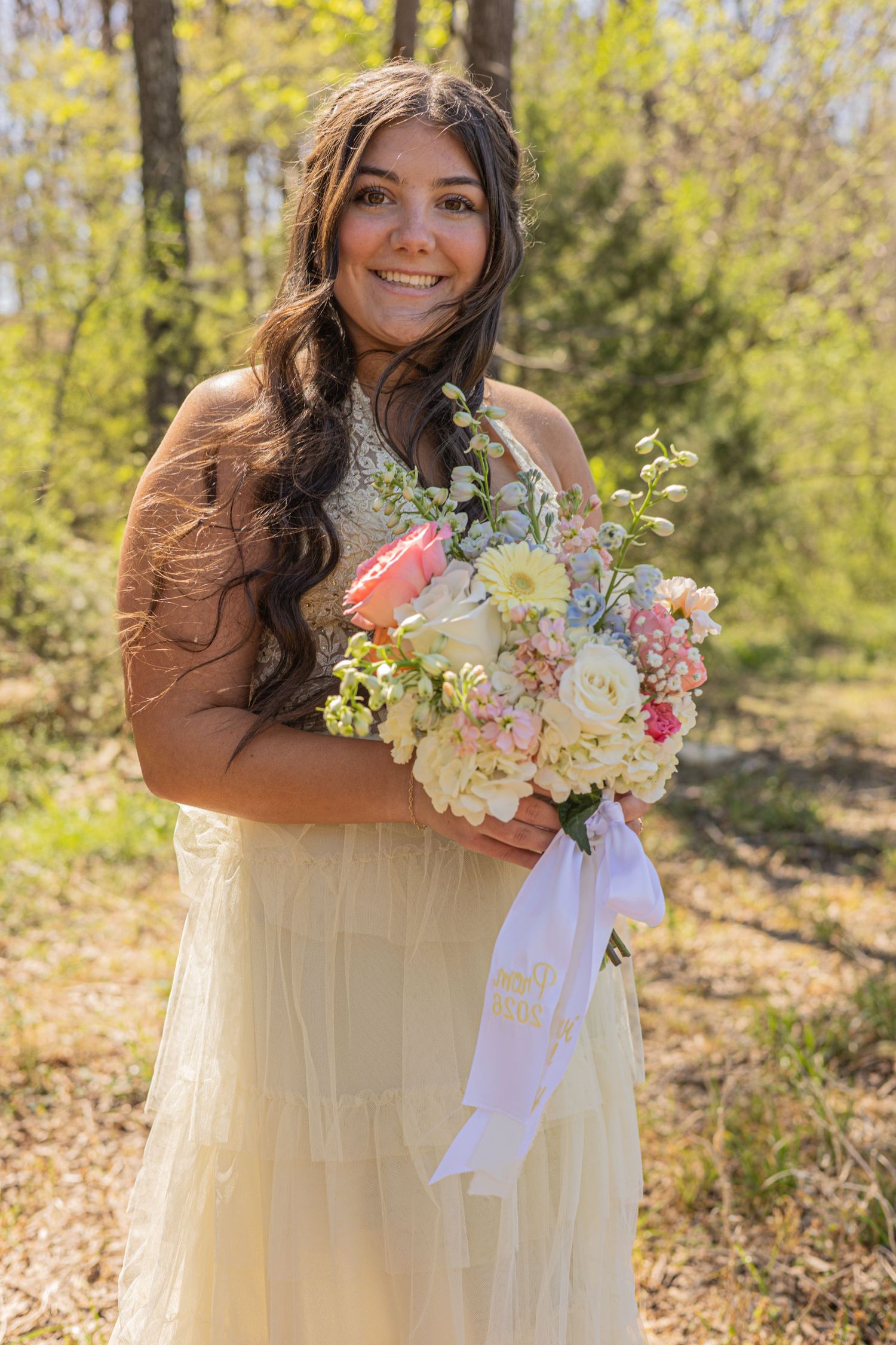 Smiling young woman in cream dress holding pink and white floral bouquet outdoors in sunlit forest setting.